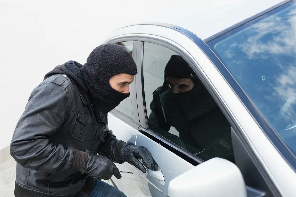 A car thief wearing a balaclava attempts to pry open a silver car's door with a tool.